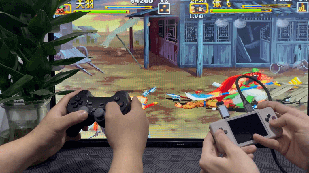 28xx-video-multiplayer - Retro Handhelds A promo photo showing two people playing an arcade game on a television. On the right, a person is playing with the RG28XX, which is plugged into the television, outputting the gameplay seen in the background. On the left, a person is playing with a generic wireless controller, which has been plugged into the RG28XX with a dongle and adapter.